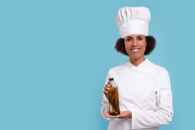 Photo of Happy female chef in uniform holding bottle of oil on light blue background. Space for text