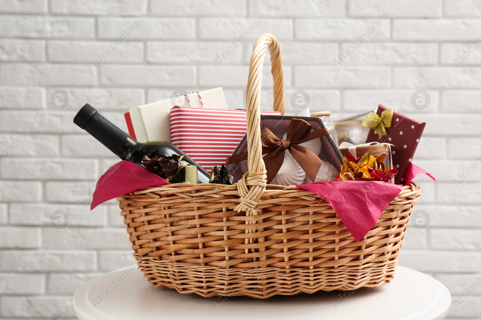 Photo of Wicker gift basket with bottle of wine on table near white brick wall