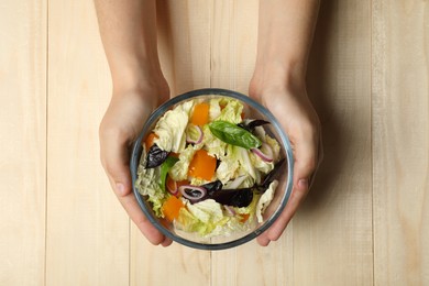 Woman holding bowl of delicious salad with Chinese cabbage, tomato and basil at wooden table, top view