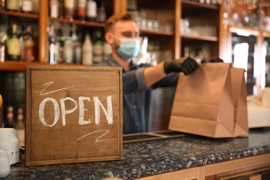 Photo of OPEN sign and blurred view of waiter with takeout orders on background. Food service during coronavirus quarantine