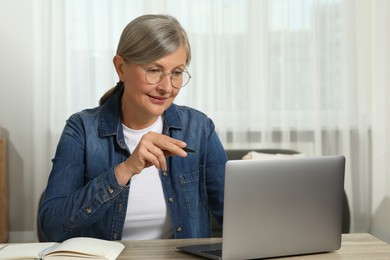 Beautiful senior woman with pen using laptop at wooden table indoors