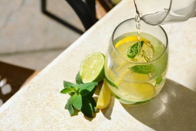 Photo of Pouring water into glass with lemon slices and mint from jug at light grey table, closeup. Space for text
