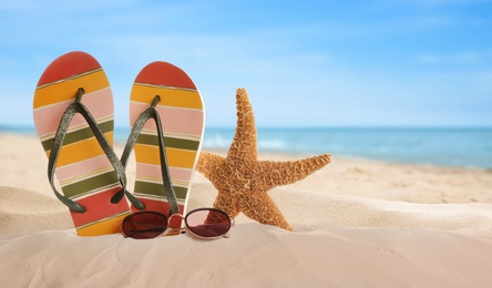 Flip flops and stylish sunglasses on sand near ocean, space for text. Beach accessories