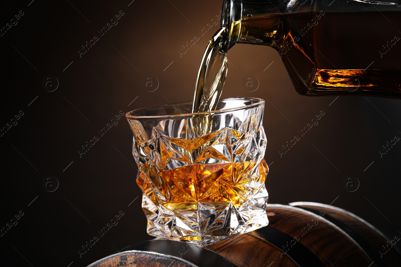 Photo of Pouring whiskey from bottle into glass on wooden barrel against black background, closeup