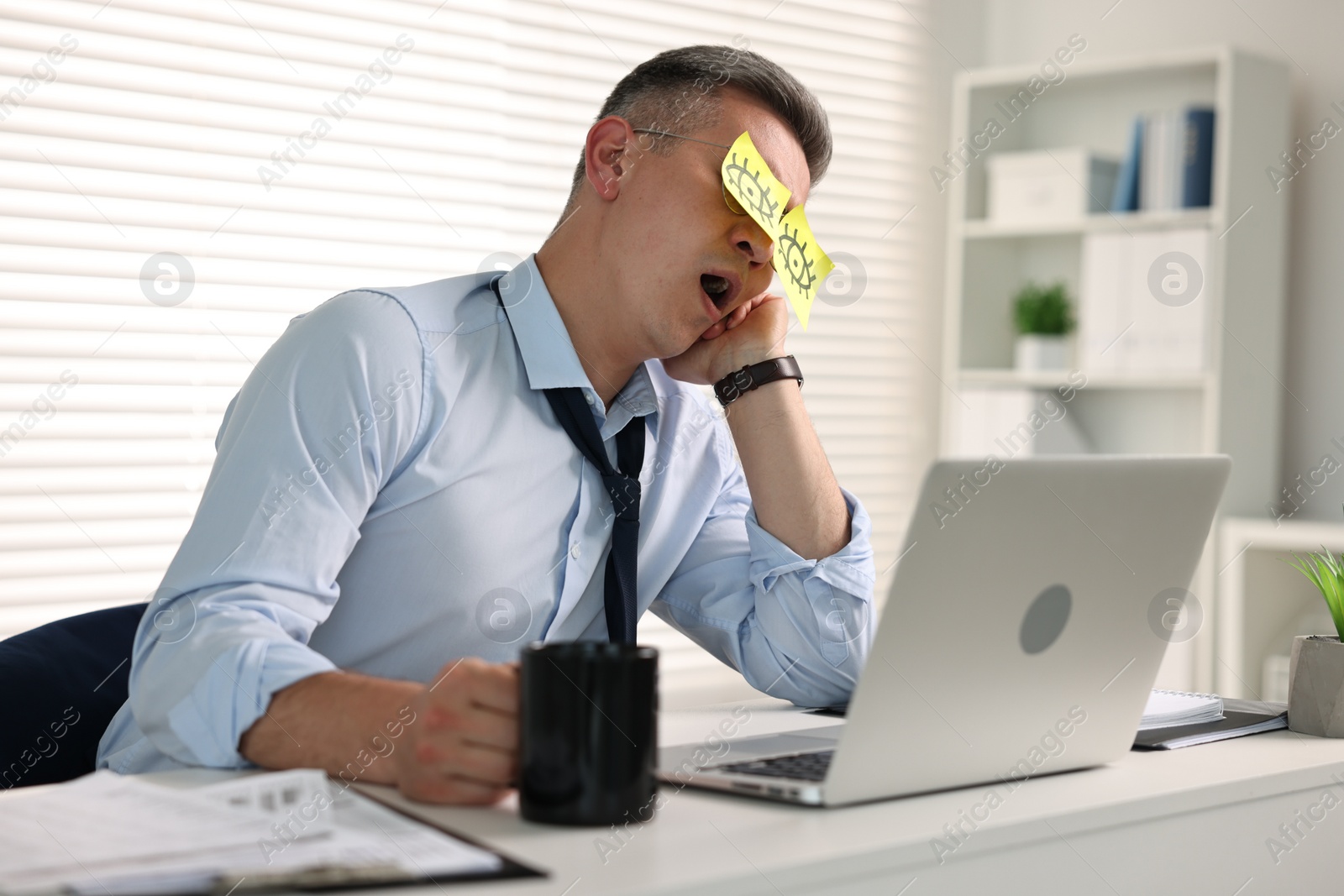 Photo of Man with fake eyes painted on sticky notes yawning at workplace in office