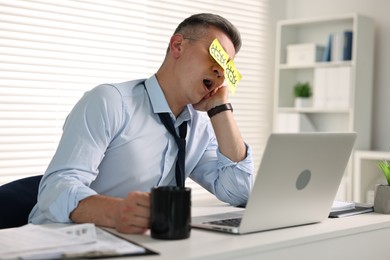 Man with fake eyes painted on sticky notes yawning at workplace in office