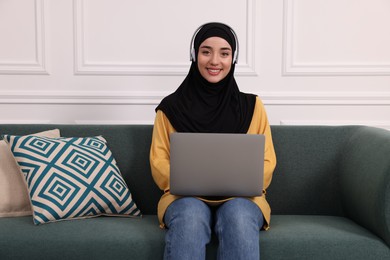 Muslim woman in hijab and headphones using laptop on sofa indoors