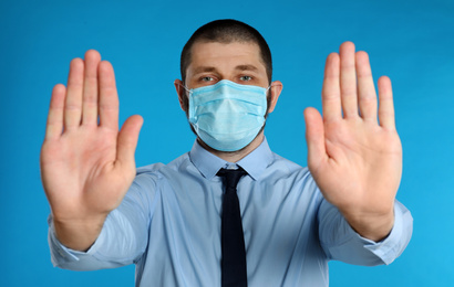 Photo of Man in protective mask showing stop gesture on light blue background. Prevent spreading of coronavirus