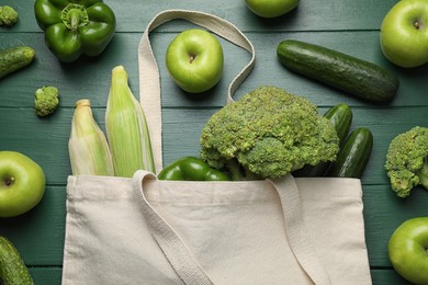 Photo of Cloth bag with fresh vegetables and apples on green wooden table, flat lay