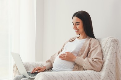 Pregnant young woman with laptop at home, space for text