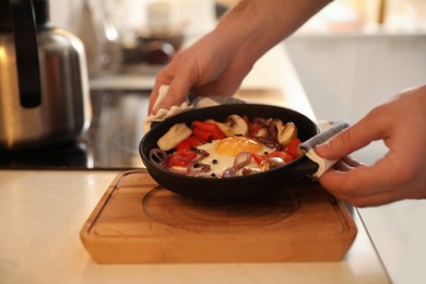 Man putting frying pan with fresh egg and vegetables on wooden board, closeup
