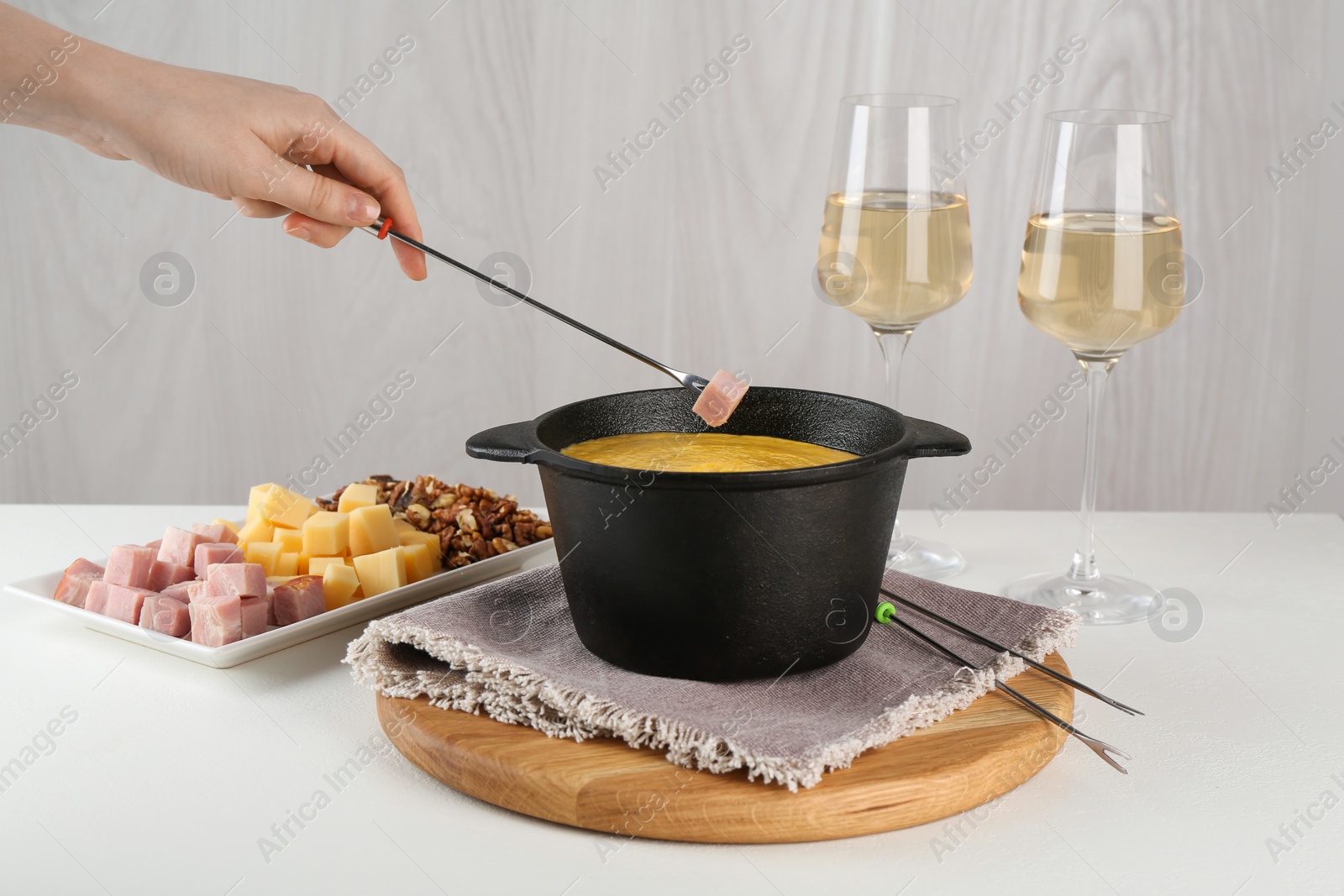 Photo of Dipping piece of ham into fondue pot with tasty melted cheese at white table, closeup