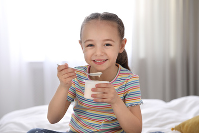 Photo of Cute little girl eating tasty yogurt on bed at home