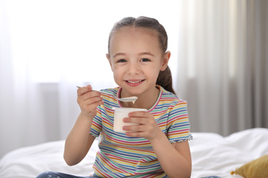 Cute little girl eating tasty yogurt on bed at home