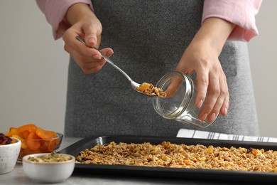 Photo of Woman putting granola from baking tray into jar at grey table, closeup