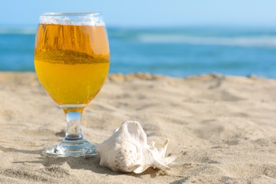 Glass of cold beer and seashell on sandy beach near sea, closeup. Space for text
