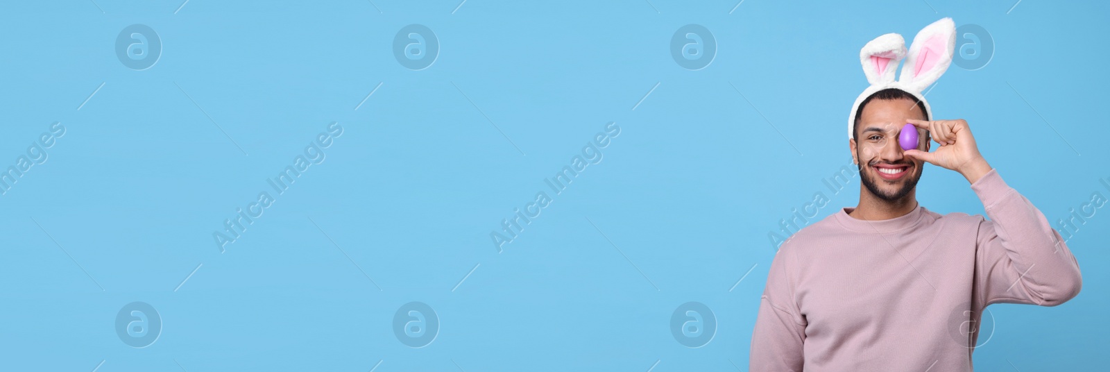 Image of Happy man with bunny ears holding Easter egg near eye on light blue background, space for text. Banner design