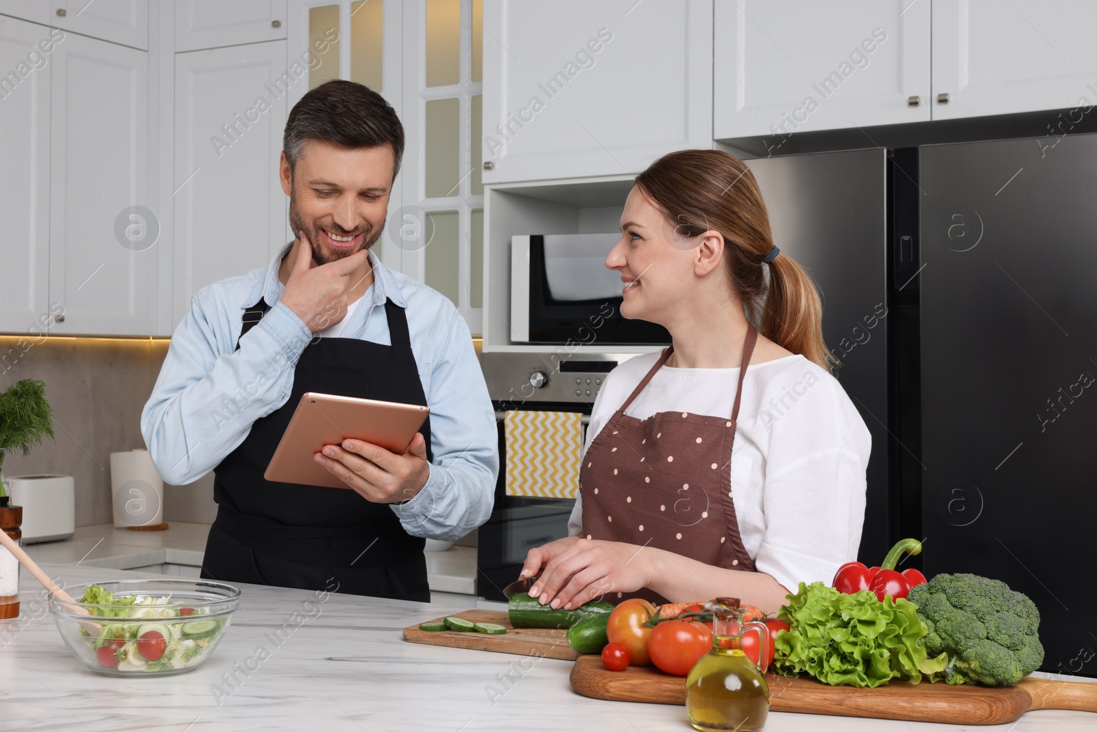 Photo of Happy couple reading recipe on tablet while cooking in kitchen. Online culinary book