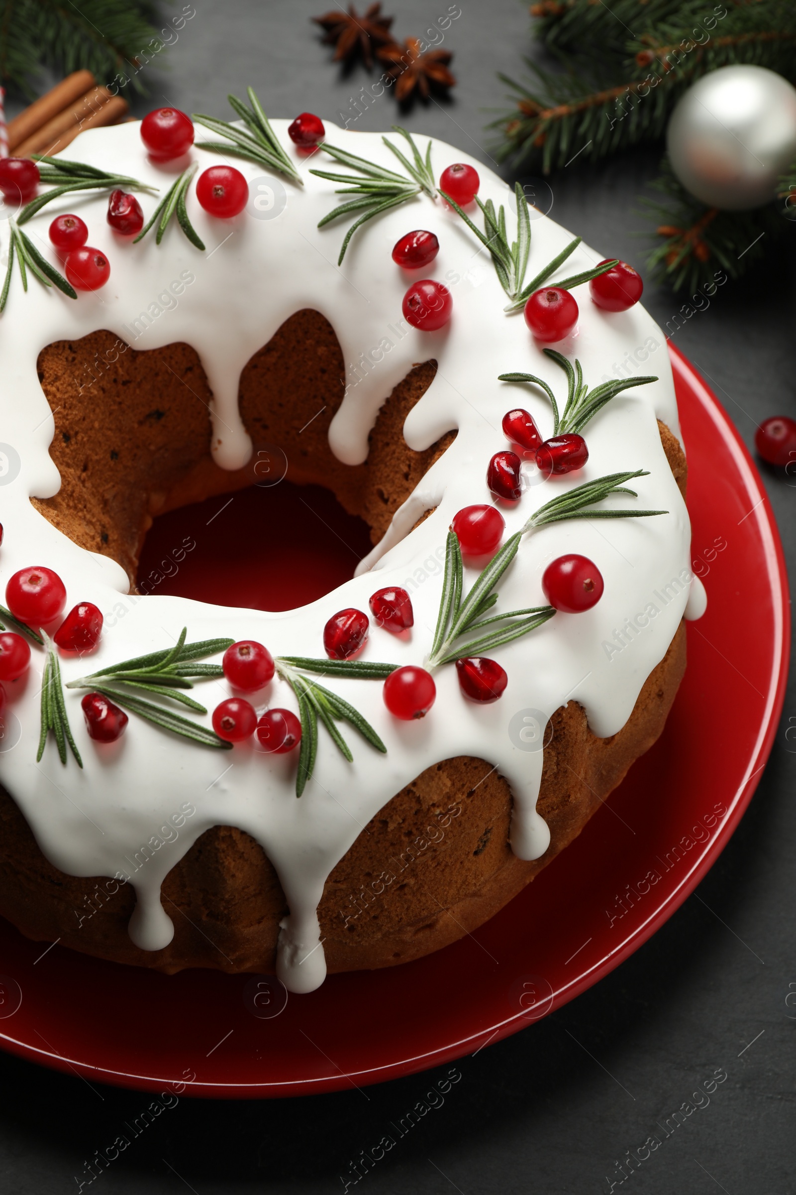 Photo of Traditional Christmas cake decorated with glaze, pomegranate seeds, cranberries and rosemary on dark grey table, closeup