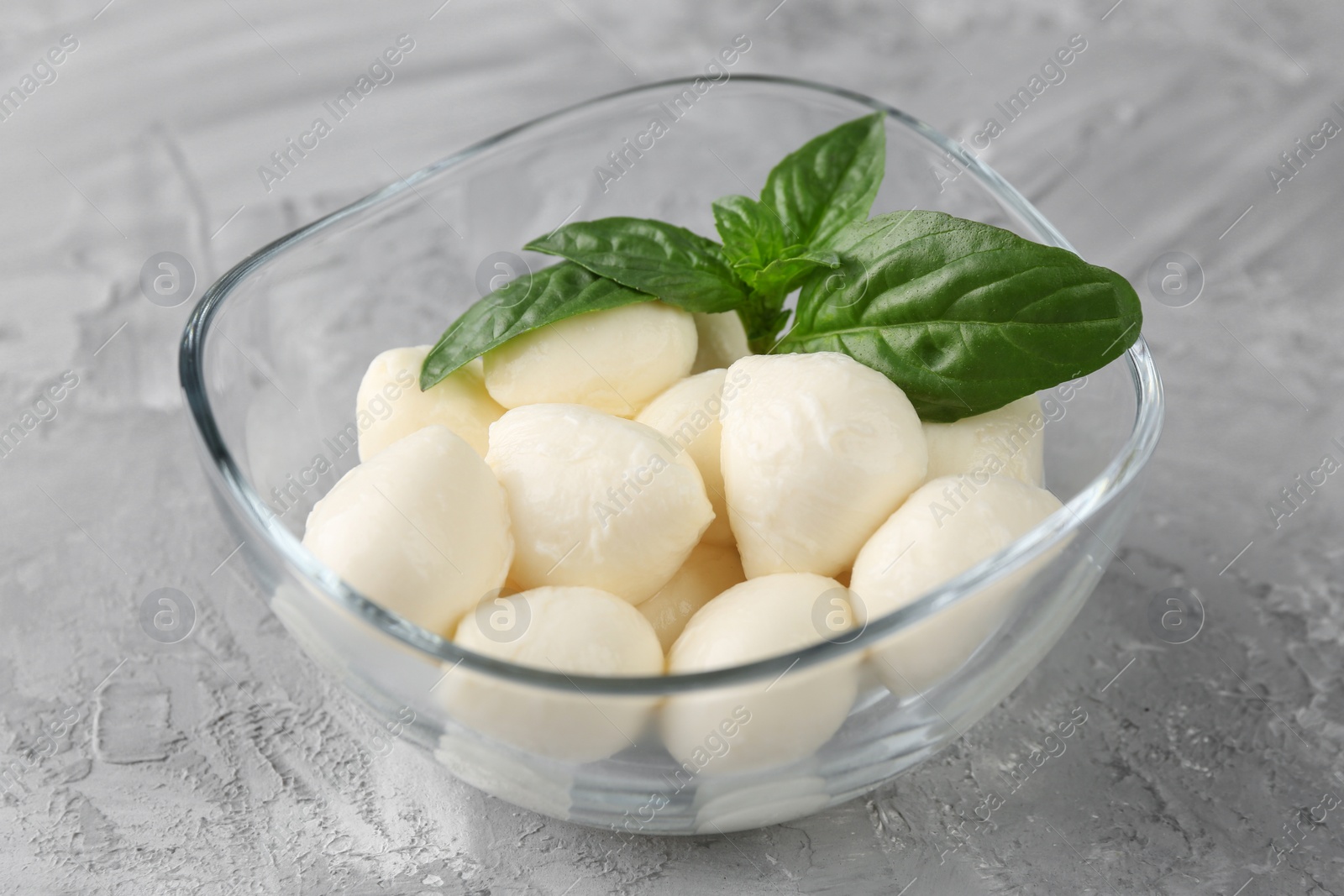 Photo of Tasty mozarella balls and basil leaves in glass bowl on grey textured table, closeup