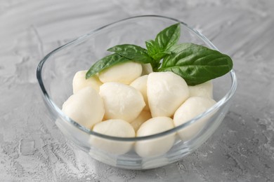 Tasty mozarella balls and basil leaves in glass bowl on grey textured table, closeup
