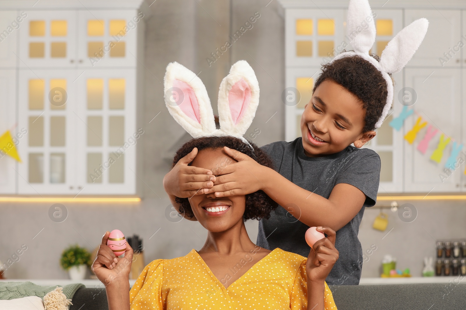 Photo of Cute African American son covering his mother's eyes with hands while she holding Easter eggs in kitchen