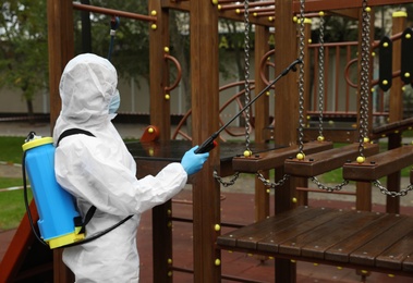 Woman wearing chemical protective suit with disinfectant sprayer on playground. Preventive measure during coronavirus pandemic