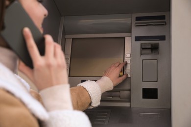 Young woman talking by mobile phone while using cash machine for money withdrawal outdoors, closeup