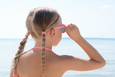 Photo of Little girl wearing sunglasses at beach on sunny day, back view