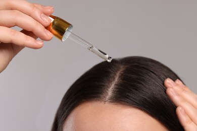 Photo of Woman applying essential oil onto hair roots on light grey background, closeup