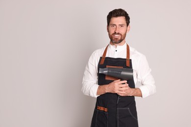 Photo of Smiling hairdresser wearing apron with dryer on light grey background, space for text