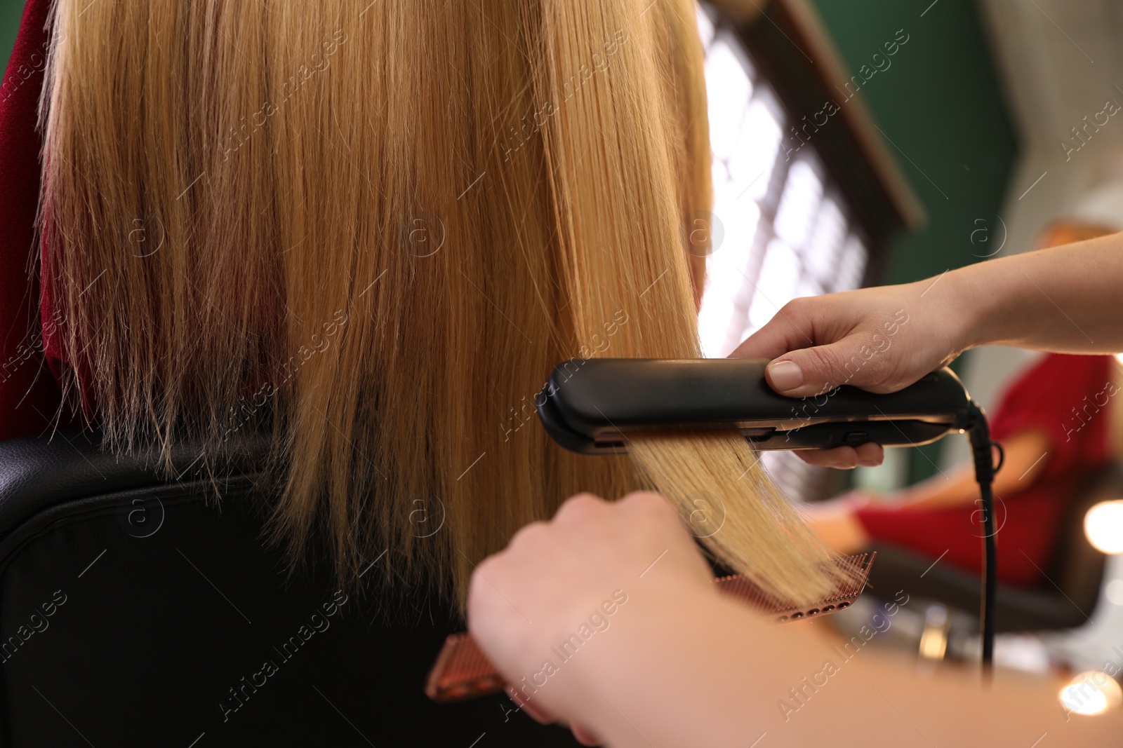 Photo of Stylist straightening woman's hair with flat iron in salon, closeup