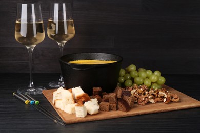 Photo of Fondue pot with melted cheese, glasses of wine and different products on black wooden table
