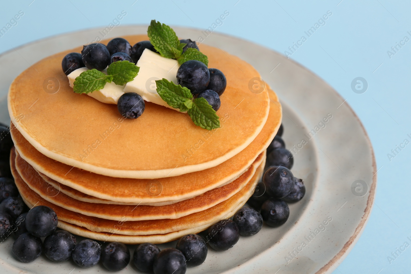 Photo of Stack of tasty pancakes with blueberries, butter and mint on light blue background, closeup