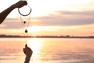 Photo of Young woman holding amulet near river at sunset, closeup view with space for text. Nature healing power