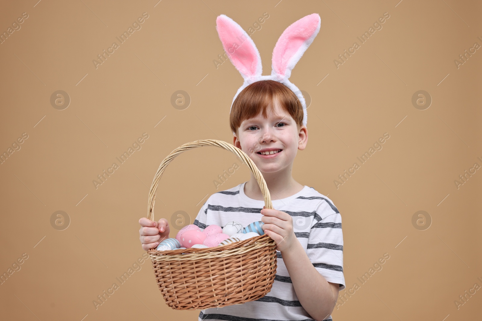 Photo of Easter celebration. Cute little boy with bunny ears and wicker basket full of painted eggs on dark beige background