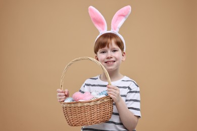 Easter celebration. Cute little boy with bunny ears and wicker basket full of painted eggs on dark beige background