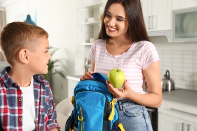 Photo of Happy mother giving apple to little child's with school bag in kitchen