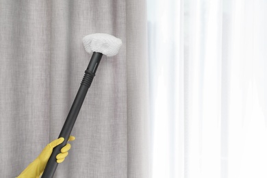 Photo of Janitor removing dust from curtain with steam cleaner indoors, closeup