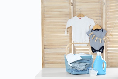 Photo of Basket with baby laundry, bottle of detergent and washing powder on white table