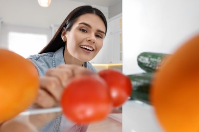 Young woman taking tomato out of refrigerator in kitchen, view from inside