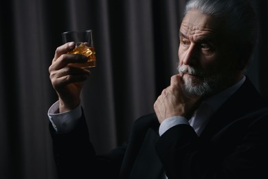 Senior man in formal suit holding glass of whiskey with ice cubes on brown background