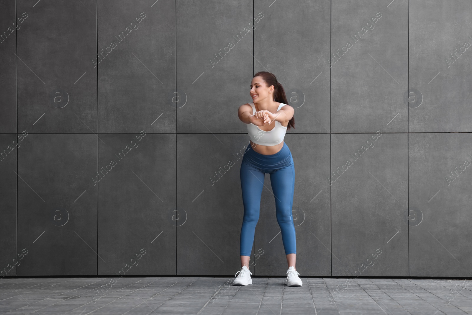 Photo of Smiling woman in sportswear stretching near dark grey wall outdoors