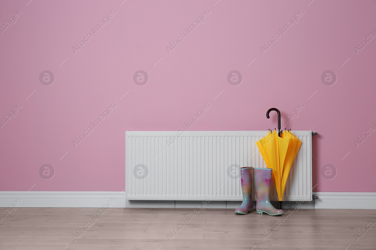 Photo of Modern radiator, rubber boots and umbrella near color wall with space for text. Central heating system