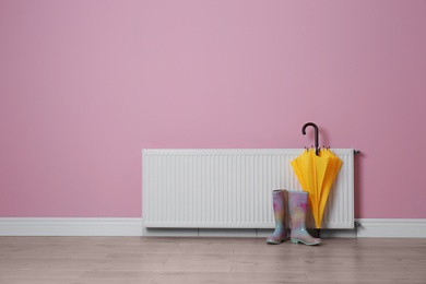 Modern radiator, rubber boots and umbrella near color wall with space for text. Central heating system