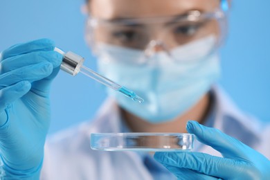 Scientist dripping liquid from pipette into petri dish on light blue background, focus on hands
