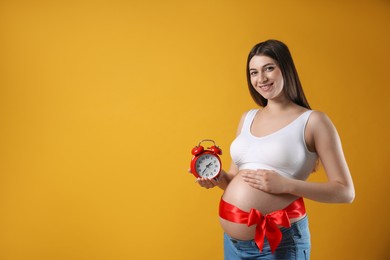 Young pregnant woman with alarm clock and red bow on yellow background, space for text. Time to give birth