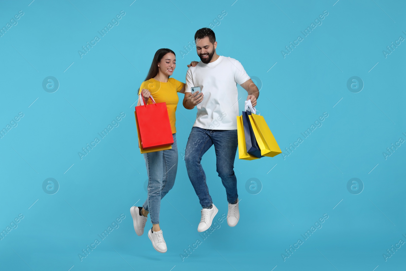 Photo of Happy couple with shopping bags and smartphone jumping on light blue background