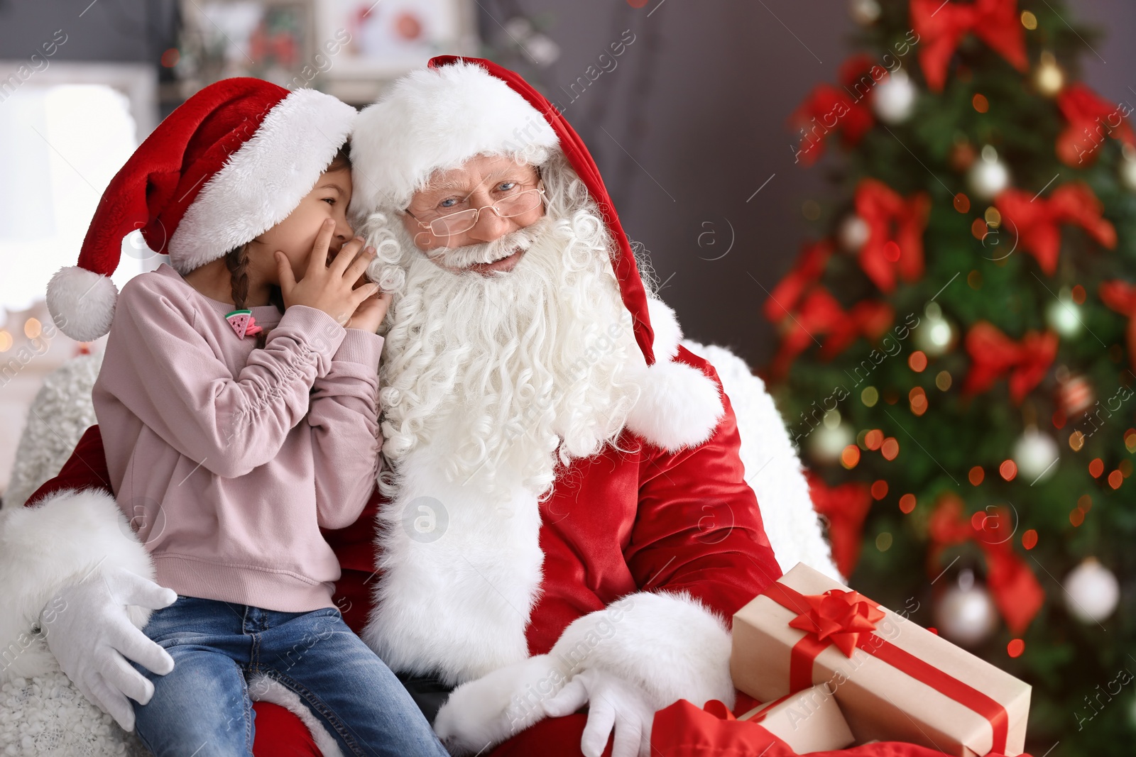 Photo of Little girl whispering in authentic Santa Claus' ear indoors