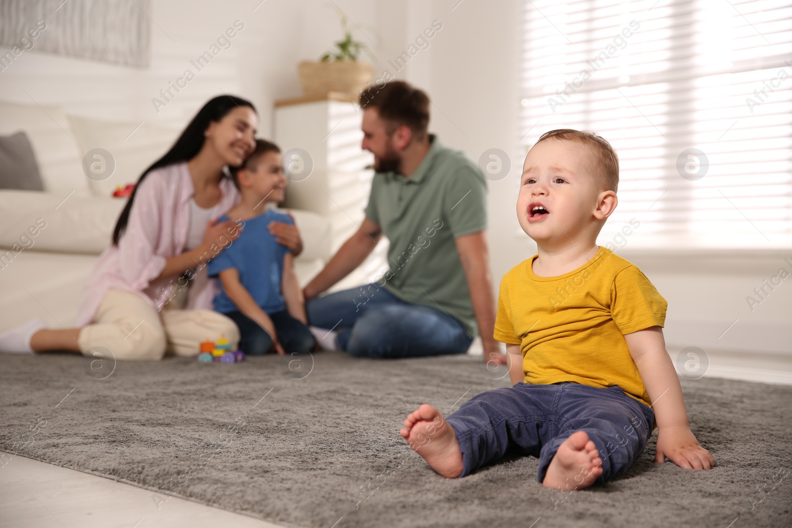 Photo of Unhappy baby sitting alone on floor while parents spending time with his elder brother at home. Jealousy in family